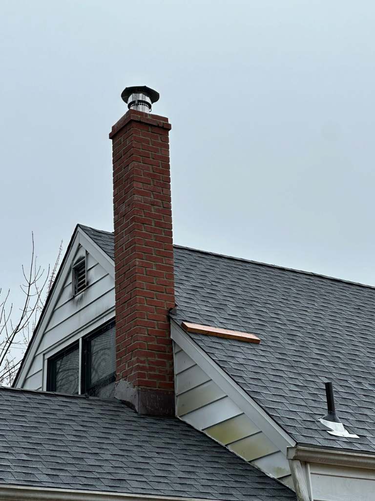 Brick chimney rising above a grey shingle roof with metal flashing at its base