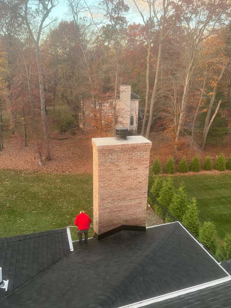 Tall brick chimney rising from a dark shingle roof, with a worker in a red jacket standing on the roof beside it