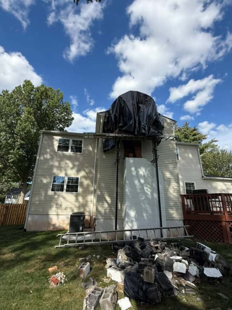 Back of a two-story house with the chimney chase partway through the work, its top under a black tarp and old material piled on the lawn below