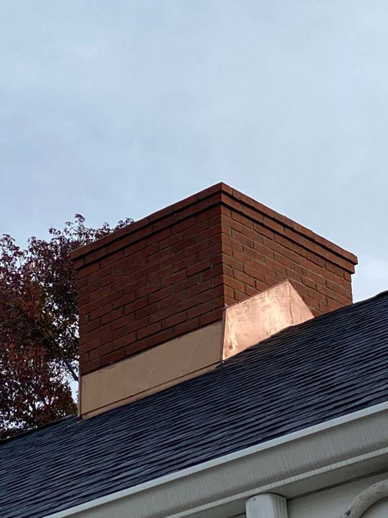 Brick chimney where it meets a dark shingle roof, with copper counterflashing along its base