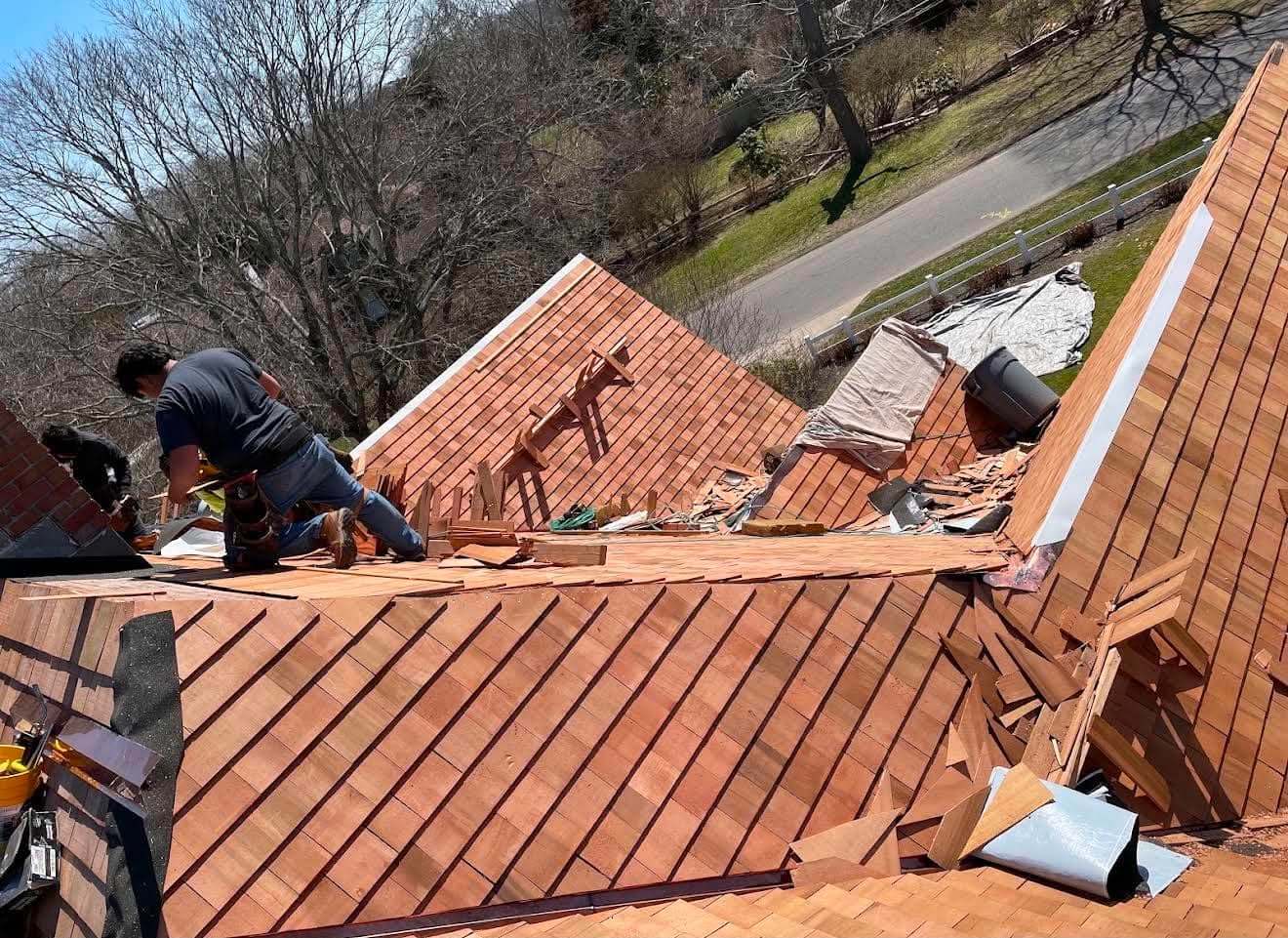 Roofer kneeling on a cedar shake roof partway through an installation, with new shakes laid across the lower slope, offcuts stacked around him and a road visible below