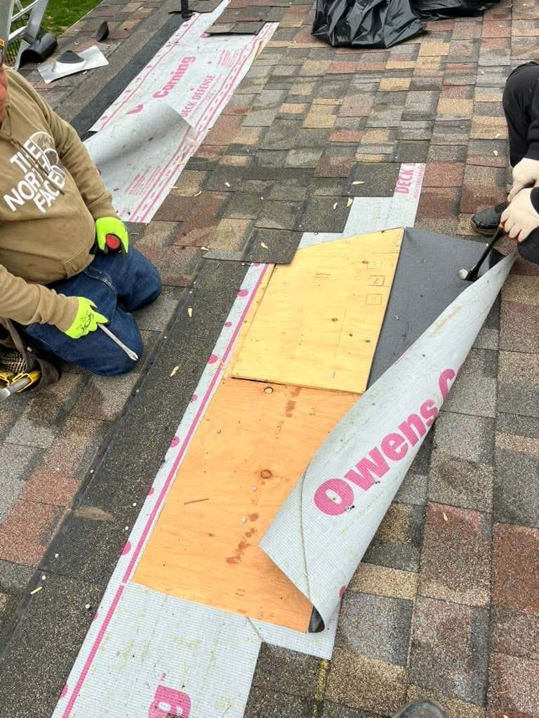 Two roofers fitting a new sheet of plywood decking into a roof, with underlayment rolled back beside them
