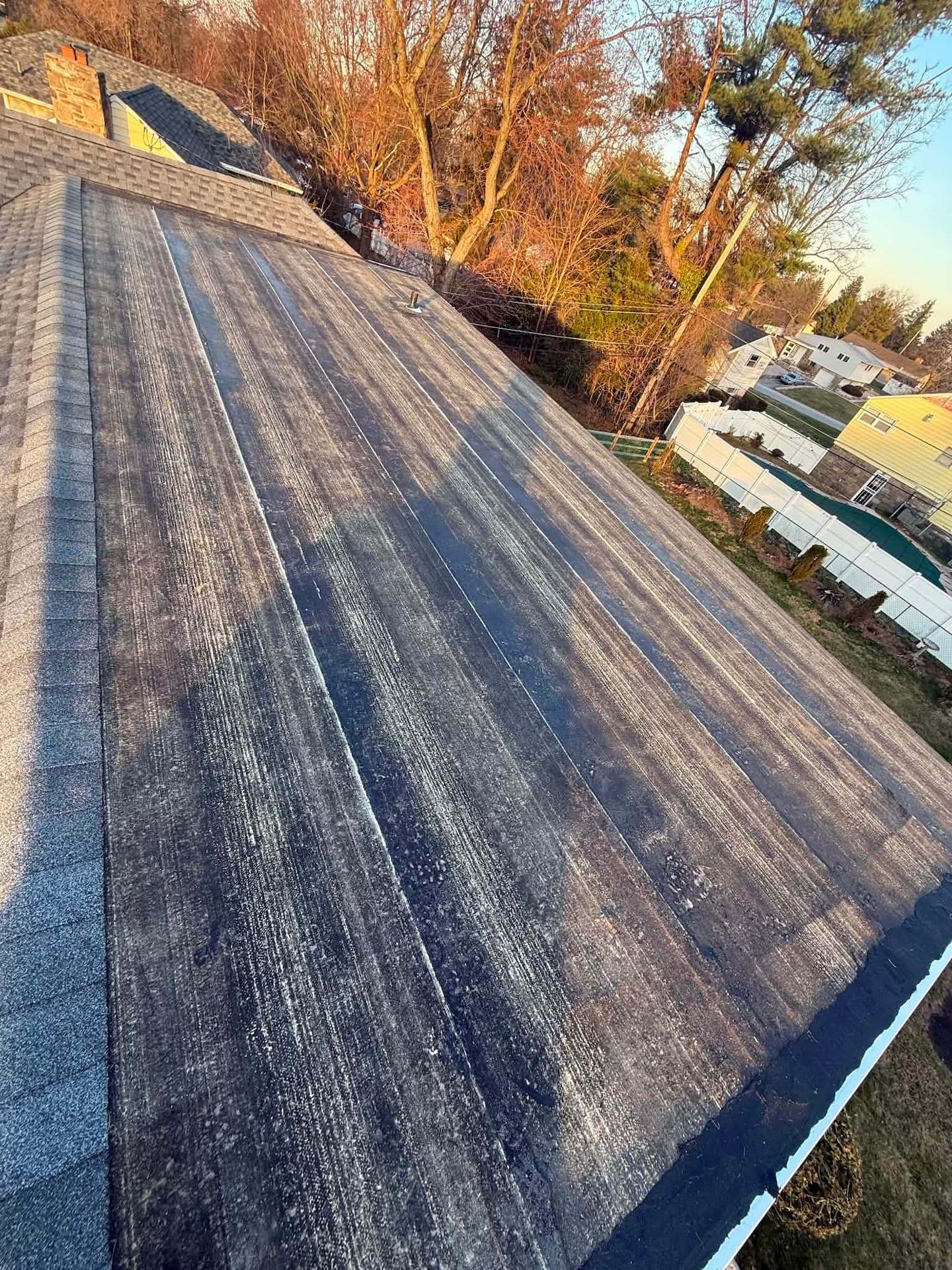 Low-slope roof section covered in dark membrane beside an asphalt shingle roof, with backyards below