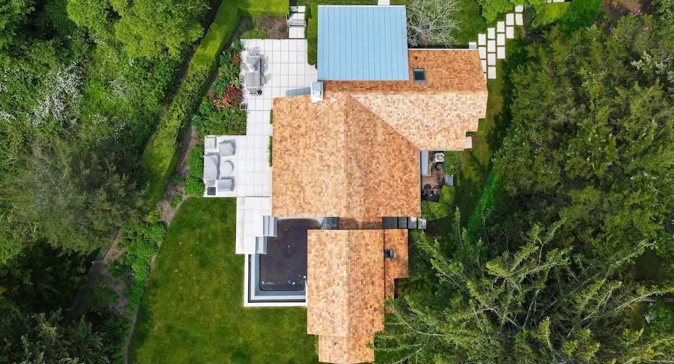 Overhead drone view of a finished cedar shake roof with a skylight and a standing seam metal section, a patio and pool beside the house
