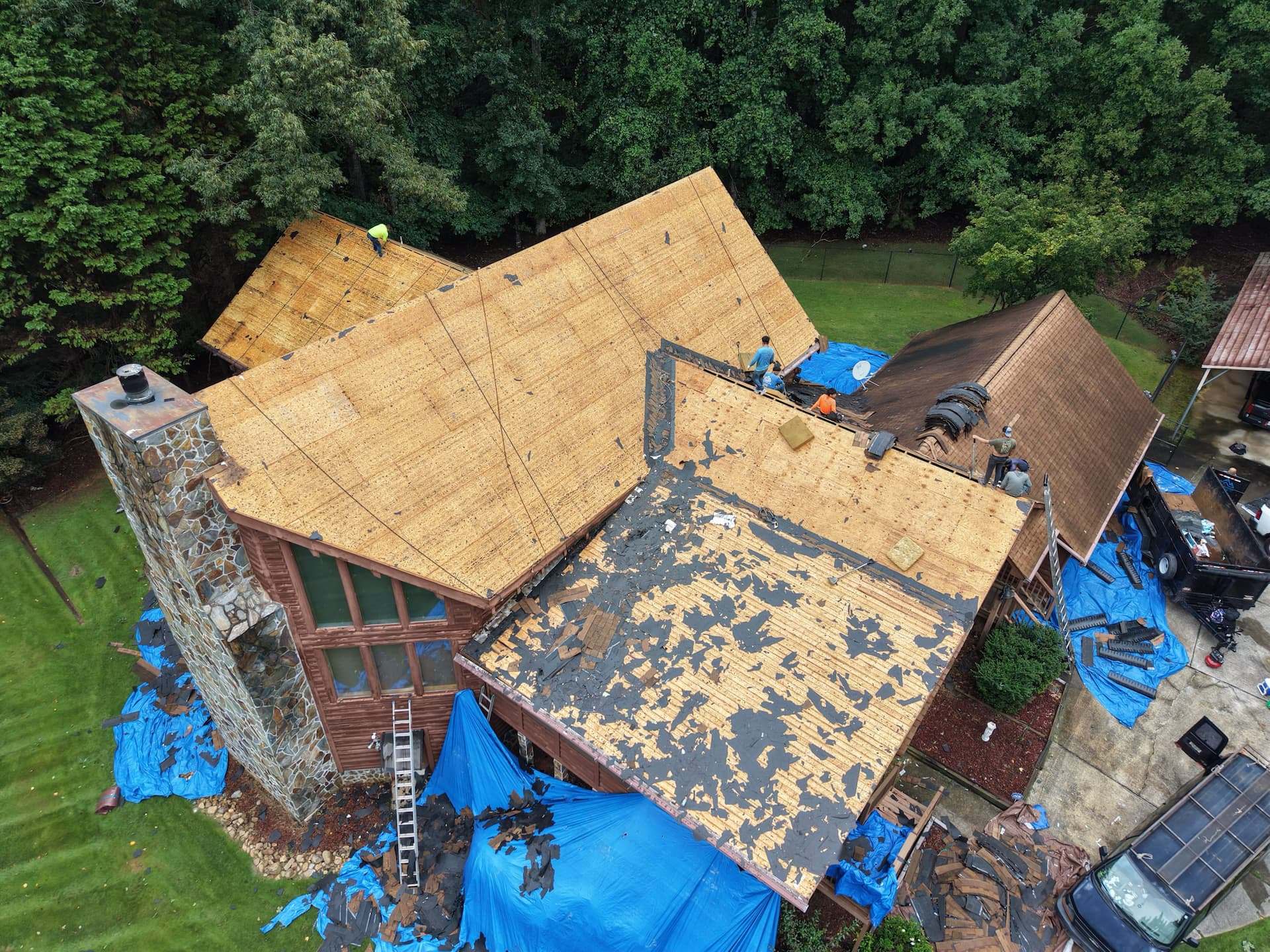 Aerial view of a house mid roof replacement, with old shingles stripped back to bare decking and blue tarps laid out on the ground