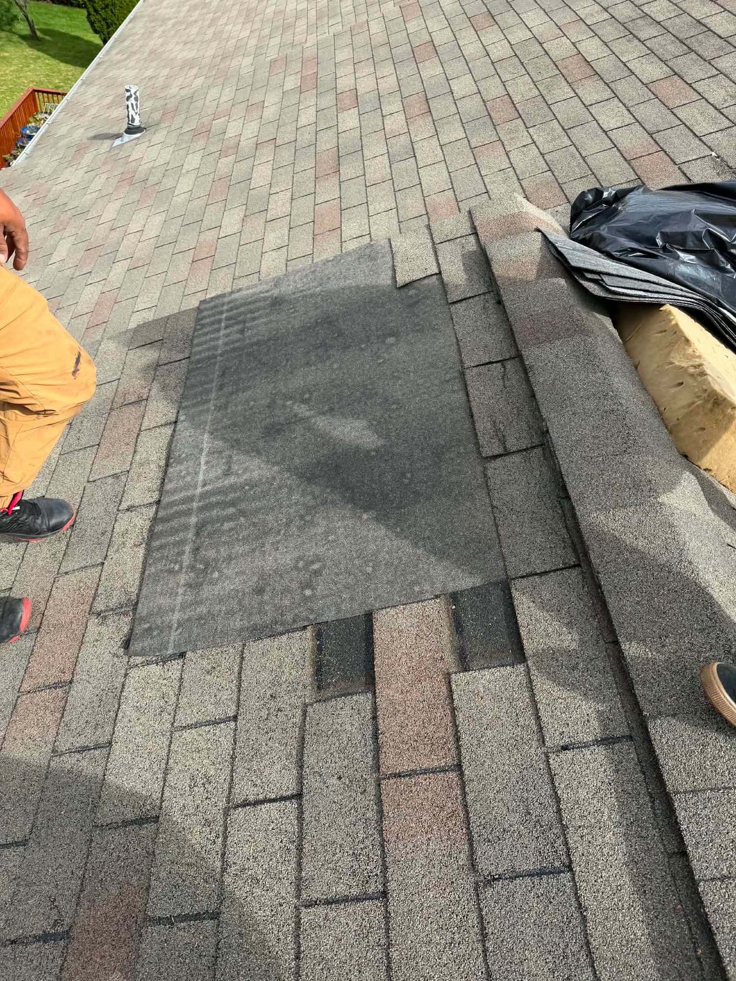 Shingles cut back on a sloped roof to expose a patch of underlayment, a roofer's boots at the edge of the frame and a bundle of replacement shingles beside the opening