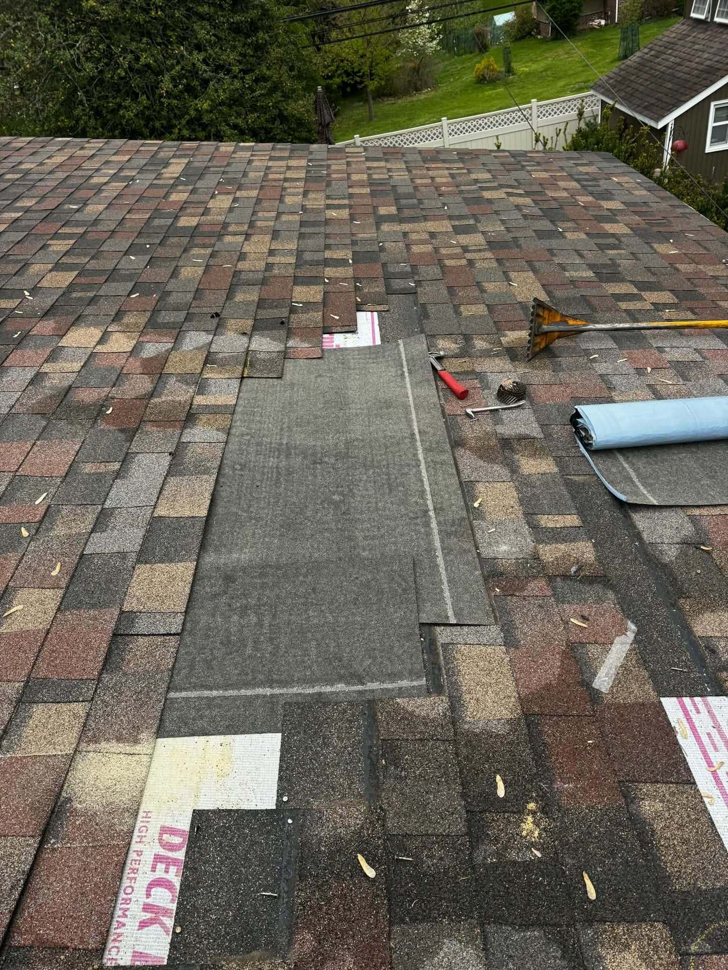 Section of an architectural shingle roof with the shingles cut back to expose the underlayment, a roofing shovel, pry bar and hammer resting beside the opening and a roll of new underlayment ready