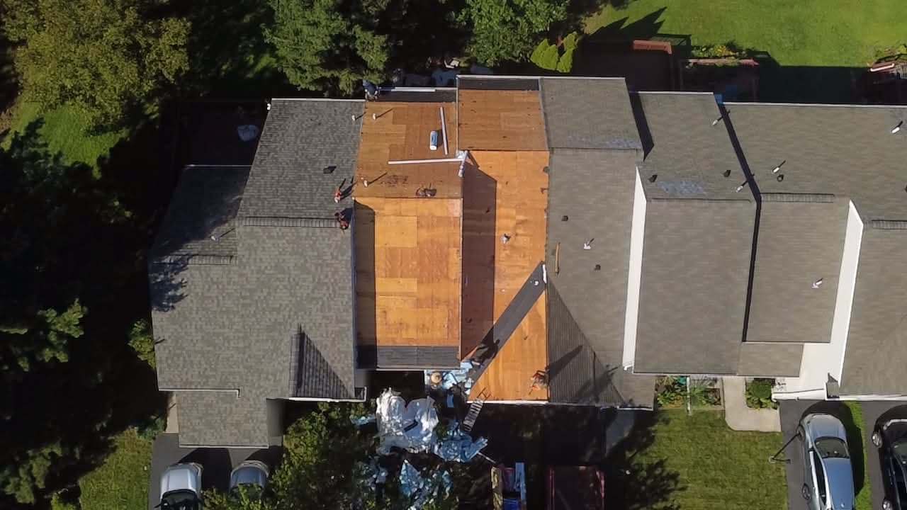 Overhead drone view of a row of attached homes, two units stripped to bare plywood decking and bagged tear-off on the ground below