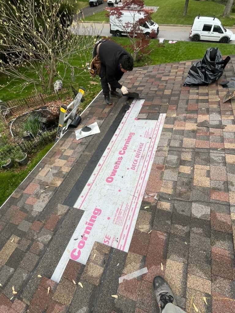 Roofer laying synthetic underlayment along a roof edge, with two work vans parked on the street below